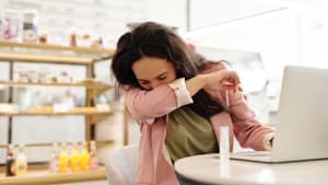Eine junge Frau sitzt am Laptop und hustet in ihre Armbeuge.