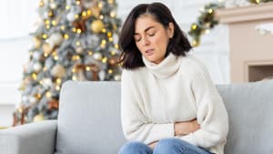 Frau im weißen Rollkrafenpullover sitzt auf der Couch und hat Bauchschmerzen