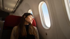 Frau mit langen, braunen Haaren sitzt im Flugzeug und schaut aus dem Fenster