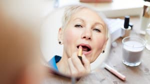 Frau mit grauen Haaren trägt vor dem Spiegel Lippenstift auf.