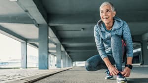 Eine Frau mit grauen kurzen Haaren und Sportkleidung kniet auf dem Boden und schnürt die Schuhe zu.