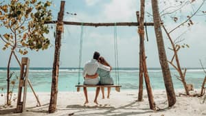 Pärchen von hinten sitzt Arm in Arm auf einer Schaukel am Strand.