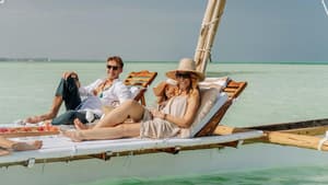 Mann und Frau mit Sonnenbrillen und sommerlicher Kleidung liegen auf einer Liege über dem Meer.