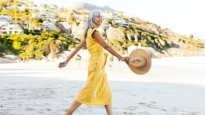 Frau mit grauen Haaren trägt ein langes, gelbes Kleid und spaziert am Strand mit einem Sonnenhut.