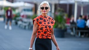 Ältere Frau mit grauen Haaren und Sonnenbrille in einem orangenen T-Shirt mit dunklem Blumenmuster.