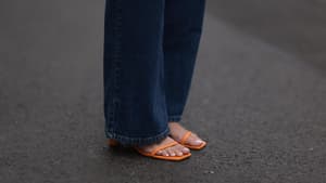 Nackte Füße in orangenen Sandalen mit hellem Nagellack und einer langen, dunklen Jeans.