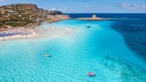 La Pelosa Strand auf Sardinien mit türkisblauem Wasser, Sonnenliegen und -schirmen und Häusern im Hintergrund.