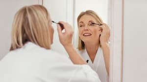 Ältere Frau mit blonden Haaren steht im Bademantel vor dem Spiegel und trägt Mascara auf.