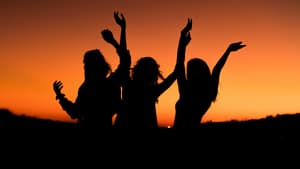 Drei Frauen tanzen ausgelassen vor dem Sonnenuntergang.