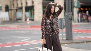 Frau mit dunkelbraunen Haaren trägt eine dunkelbraune Hose und transparente Bluse mit Muster und fässt sich mit einem Arm an den Kopf..