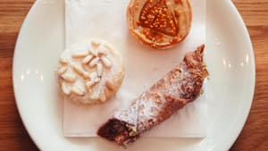 Drei italienische Backwaren (Cannoli, Torta della Nonna) auf einem weißen Teller, von oben nach unten auf einem Holztisch geschossen.