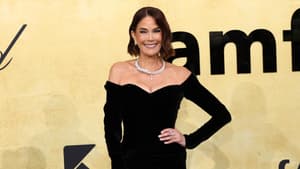 Teri Hatcher in einer schwarzen Robe mit hohem Beinausschnitt, silberner Kette und schwarzer Clutch vor einem gelben Hintergrund.