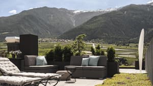 Ein Blick auf eine Terrasse mit Liegen und Sesseln und grünen Wiesen und Bergen im Hintergrund.