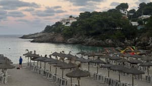 Ein Strand mit vielen Sonnenschirmen aus Stroh und Bäumen auf einem Hang im Hintergrund.
