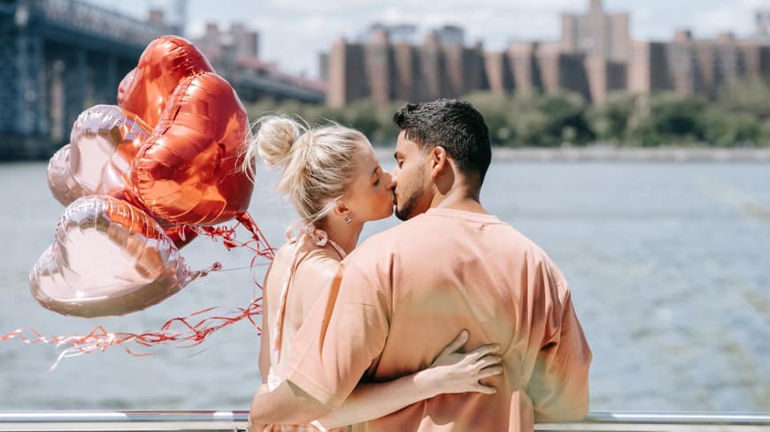 Verliebtes Paar küsst sich am Wasser und hält Herzluftballons in der Hand