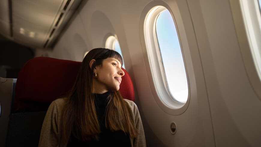 Frau mit langen, braunen Haaren sitzt im Flugzeug und schaut aus dem Fenster
