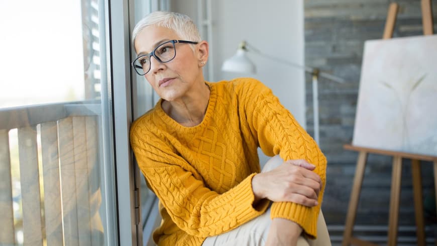 Frau mit grauen Haaren und Brille lehnt gegen das Fenster und schaut nach draußen.