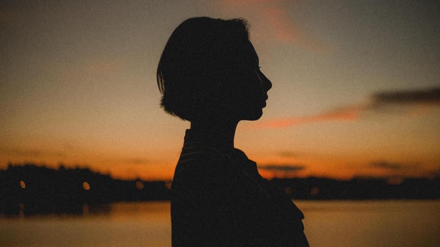 Frau steht vor dem Himmel, an dem die Sonne untergeht, und guckt zur Seite.