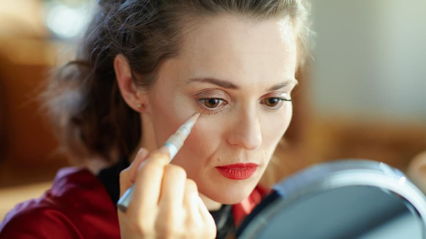 Frau sitzt vor dem Spiegel und deckt ihre Augenringe mit Concealer ab.