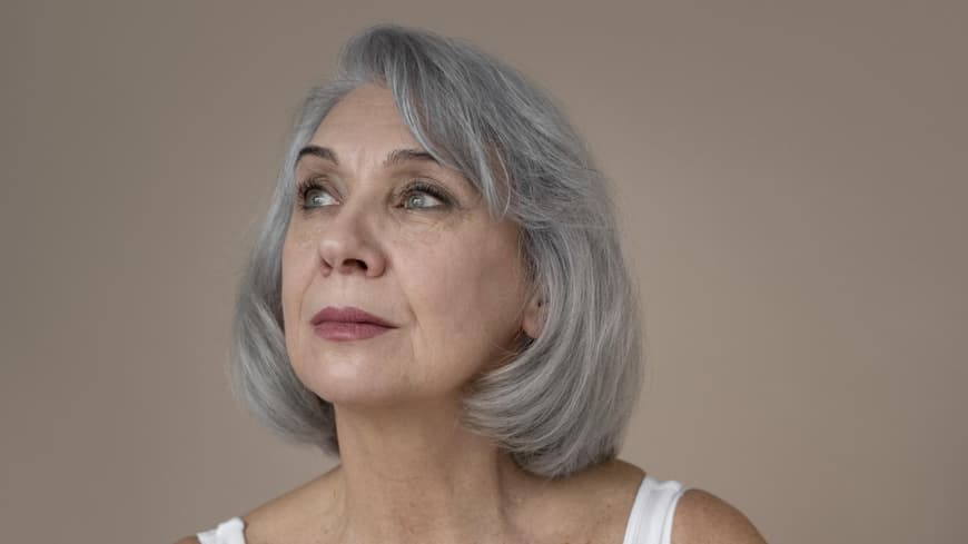 Frau mit grauen kurzen Haaren trägt eine weiße Bluse und Lippenstift.