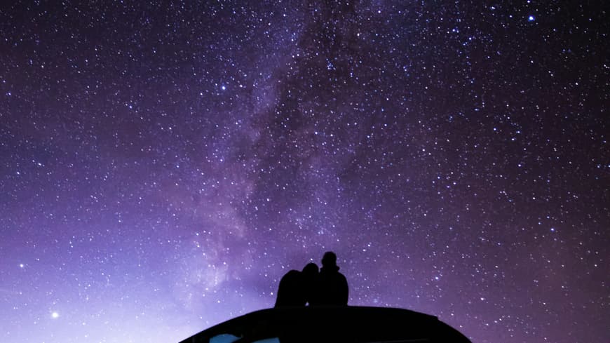 Pärchen sitzt auf Autodach und schaut in den Sternenhimmel