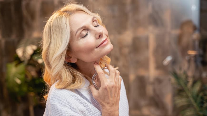 Blonde Frau trägt mit geschlossenen Augen Parfum an ihrem Hals auf und lächelt dabei.