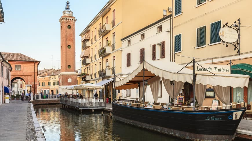 Trattorie und Häuser mit Blick auf den Kanal, der durch Comacchio fließt. Im Hintergrund plaudern Einheimische