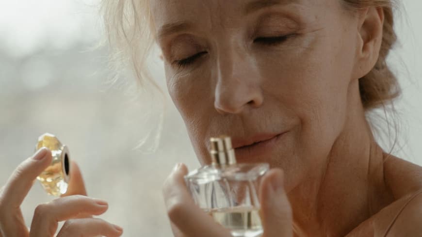 Eine ältere Frau riecht mit geschlossenen Augen an einem Parfum Flakon.