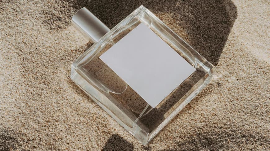 Ein Flakon liegt im Sand mit einer Muschel daneben.