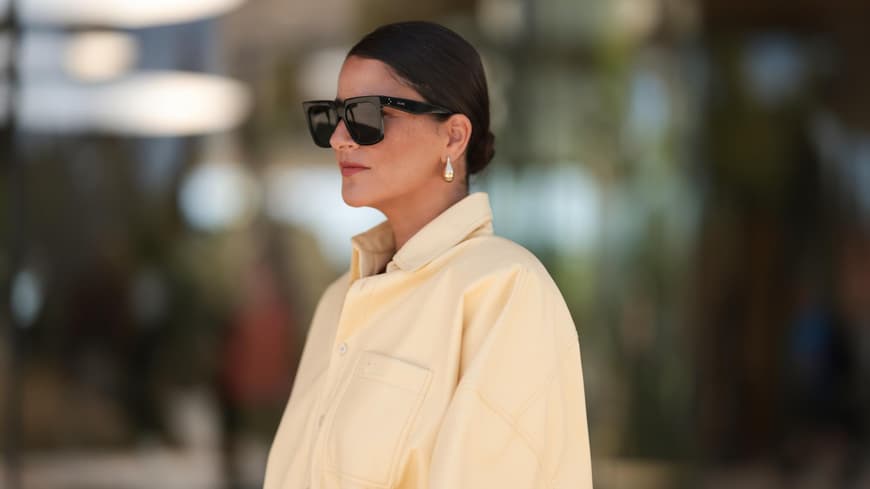 Frau mit einer gelben Jeansbluse, Sonnenbrille und einem niedrigen Dutt mit kurzen Haaren.