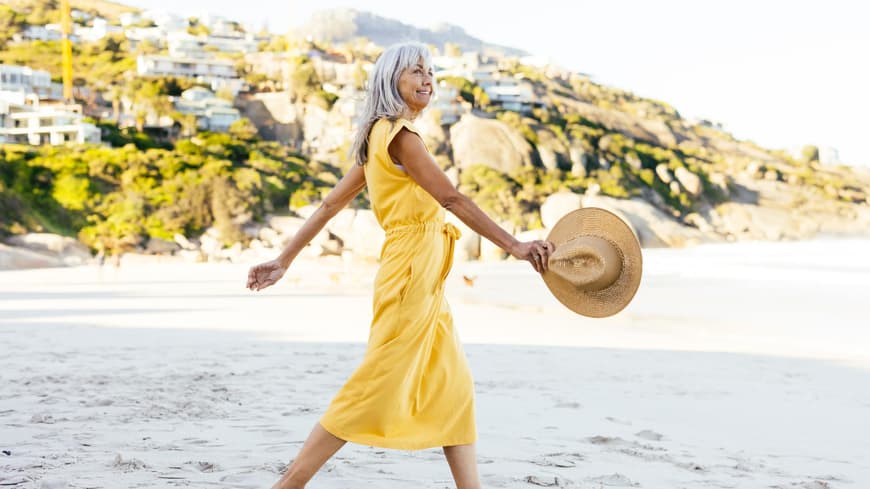Frau mit grauen Haaren trägt ein langes, gelbes Kleid und spaziert am Strand mit einem Sonnenhut.