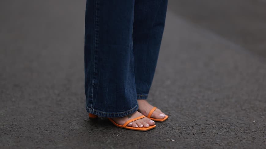 Welche Nagellackfarbe für ältere Füße geeignet ist, hängt vom Hauttyp ab. Nackte Füße in orangenen Sandalen mit hellem Nagellack und einer langen, dunklen Jeans.