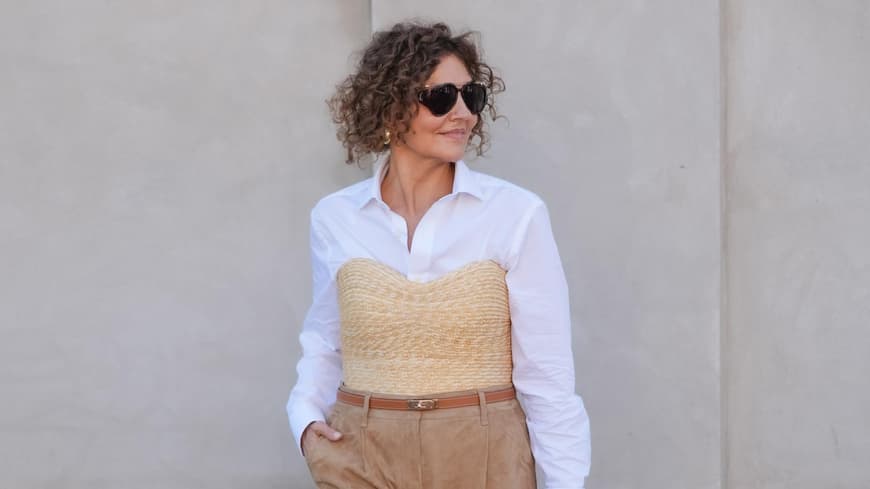 Frau mit lockigen Haaren trägt eine weiße Bluse mit einer Korb-Korsage und einer knielangen Hose.
