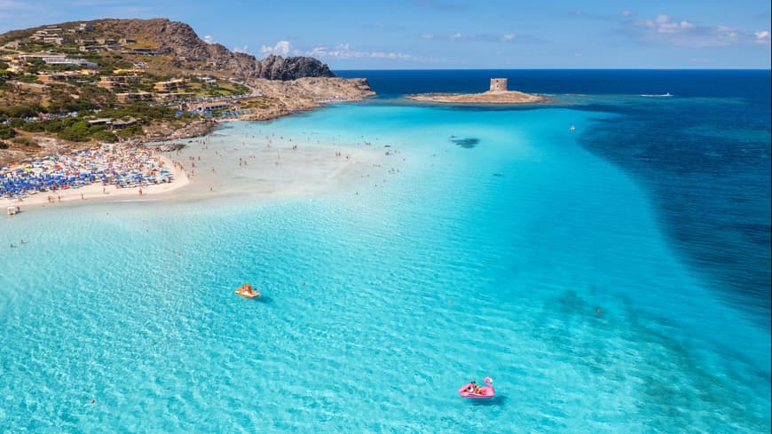La Pelosa Strand auf Sardinien mit türkisblauem Wasser, Sonnenliegen und -schirmen und Häusern im Hintergrund.