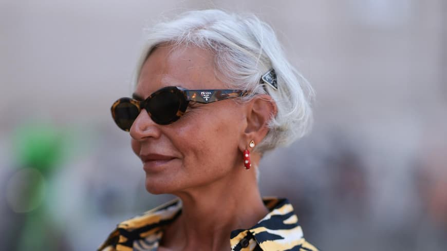 Frau mit grauen kurzen Haaren, einer Sonnenbrille und einem getigerten Oberteil guckt zur Seite.