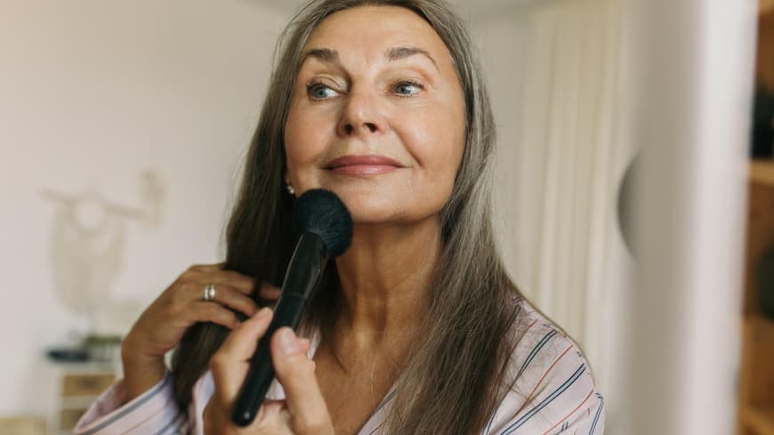 Ältere Frau mit grauen Haaren sitzt vor einem Spiegel und geht mit einem Puderpinsel über ihr Kinn.