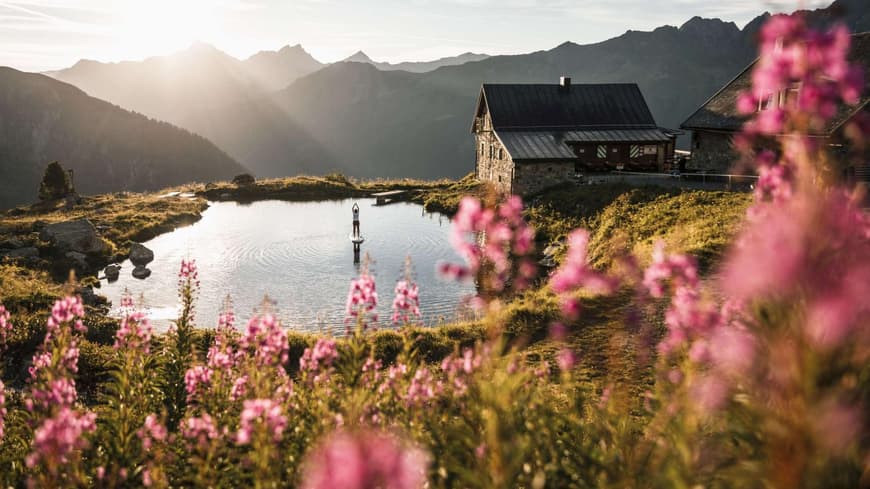 Ein Haus am See mit Gebirge im Hintergrund, rosanen Blüten im Vordergrund und Sonnenstrahlen.