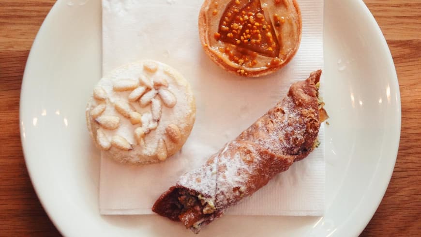 Drei italienische Backwaren (Cannoli, Torta della Nonna) auf einem weißen Teller, von oben nach unten auf einem Holztisch geschossen.