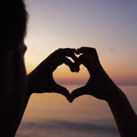 Person macht Herz mit Händen vor Sonnenuntergang