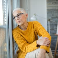 Inflammaging betrifft jeden, weshalb man sich damit auseinander setzen sollte. Frau mit kurzen grauen Haaren lehnt gegen ein Fenster und guckt nach draußen.