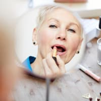 Frau mit grauen Haaren trägt vor dem Spiegel Lippenstift auf.