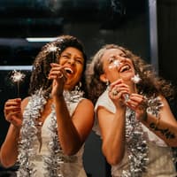 Zwei Frauen mit Wunderkerzen und Glitzerdeko um den Hals lachen.