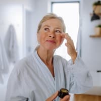 Ältere Frau trägt im Bademantel eine Creme unter ihren Augen auf.