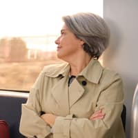 Frau mit grauen Haaren und Trenchcoat sitzt im Zug und guckt zur Seite nach draußen.