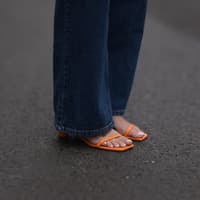 Nackte Füße in orangenen Sandalen mit hellem Nagellack und einer langen, dunklen Jeans.