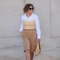 Ein Kurzhaarschnitt für lockiges Haar im Sommer ist praktisch und stylish zugleich. Frau mit lockigen Haaren trägt eine weiße Bluse mit einer Korb-Korsage und einer knielangen Hose.