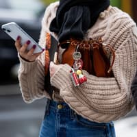 Schlüsselanhänger in allen Variationen sind Trend. Frau mit einem Cardigan hält ihre Tasche unterm Arm und ein Handy in der Hand. An der Tasche ist ein Schlüsselanhänger.