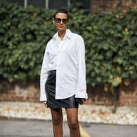 Frau mit kurzen Haaren trägt eine weiße Bluse, einen kurzen Lederrock und Loafer mit weißen Socken.