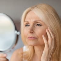 Ältere Frau schaut skeptisch in einen Handspiegel und fässt sich mit der Hand ins Gesicht.