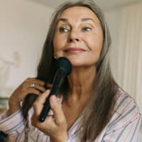 Beim Sommer-Make-up können uns einige Schminkfehler schnell älter aussehen lassen. Ältere Frau mit grauen Haaren sitzt vor einem Spiegel und geht mit einem Puderpinsel über ihr Kinn.